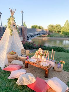 an outdoor picnic setting with pillows, plates and candles on the grass next to a river