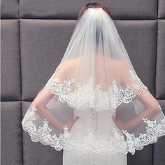 a woman wearing a wedding veil with white lace on the top and bottom, standing in front of a gray wall