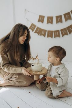 a little boy sitting on the floor next to a woman with a cake in front of him