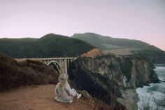 a woman sitting on top of a cliff next to the ocean with a bridge in the background