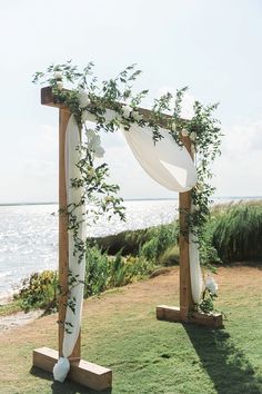 an outdoor ceremony setup with white draping and greenery on the grass by the water