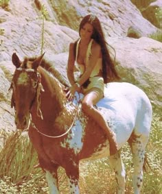 a woman sitting on top of a brown and white horse next to a rocky hillside