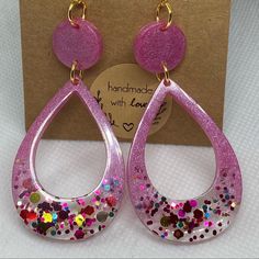 a pair of pink glittered earrings with hearts on the front and bottom, sitting next to a cardboard box