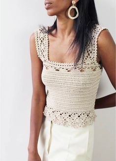 a woman wearing a crochet top with large hoop earrings standing against a white wall