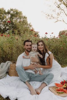 a man and woman sitting on a blanket with a baby in their lap, surrounded by flowers