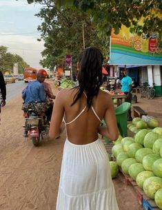 a woman in a white dress walking down a street next to watermelon stands