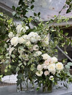 two vases filled with white flowers and greenery on a table in a greenhouse