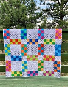 a multicolored quilt hanging on a wooden fence in front of some pine trees