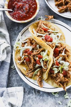 three tacos with shredded meat, tomatoes and cabbage on plates next to chili sauce