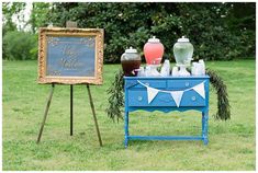 a blue table topped with vases and an easel filled with bottles on top of green grass