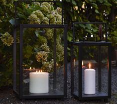 two black lanterns with white candles sitting on gravel in front of trees and bushes at night