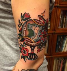 a woman's arm with a globe and flowers tattoo on her left arm, in front of a bookshelf