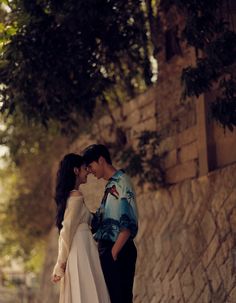 a man and woman standing next to each other near a stone wall with trees in the background