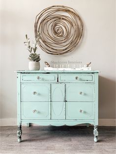 a blue dresser with two drawers and a plant on top, in front of a circular wall decoration