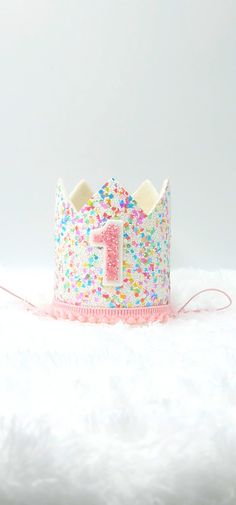 This fun Confetti crown is so sweet. This is a great addition to any celebration or cake smash photos. It is made with adorable non shedding white sprinkle glitter, white felt lining and soft pink pom trim. This crown stands 3.5 inches tall with a 3 inch diameter. The elastic is meant to be worn behind the head and not under the chin to help make it more comfortable. Whimsical White Party Supplies For Birthday, Whimsical White Birthday Party Supplies, Whimsical Tall Crown For Birthday, Funfetti Smash Cake First Birthdays, Playful Pink Tops For First Birthday, Cute Birthday Crown With Round Shape, Fabric Birthday Crown, Princess Crown Party Supplies For Birthday, Whimsical Pink Crown For Birthday