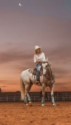 a woman is sitting on top of a white horse in an arena at sunset with the moon behind her