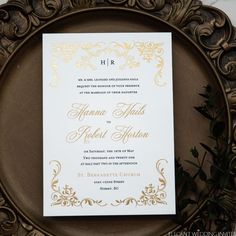 an ornate gold and white wedding card on a metal plate with greenery around it