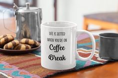 a white coffee mug sitting on top of a wooden table next to two plates of cookies