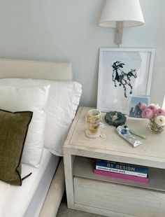 a nightstand with books and flowers on it next to a white bed in a bedroom