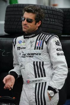 a man standing next to a pile of tires in a race suit and holding a steering wheel