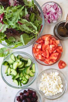 several bowls filled with different types of vegetables on top of a white counter next to olives, tomatoes, cucumbers and lettuce