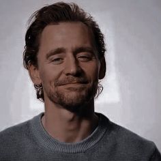 a man with long hair and beard wearing a gray sweater smiles at the camera while standing in front of a white background