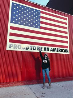 a woman holding up a sign that says proud to be an american on the side of a building