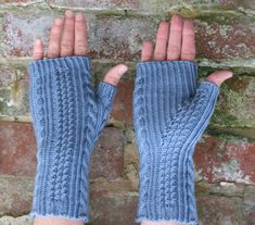 a woman's hands wearing blue knitted mitts against a brick wall with her fingers in the air