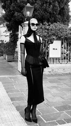 a woman in black dress and sunglasses standing on sidewalk next to lamp post with trees behind her