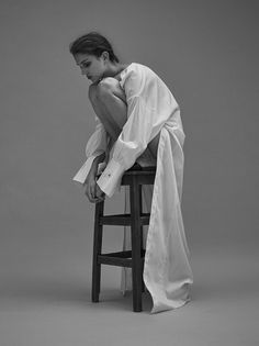 a woman sitting on top of a wooden stool in white clothing with her legs crossed