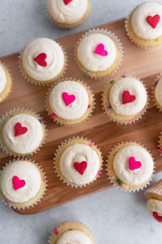 cupcakes with white frosting and red heart decorations on a wooden board next to other cupcakes