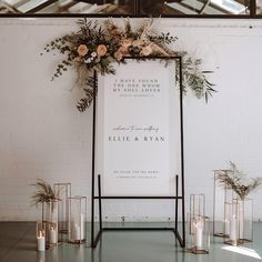 a wedding sign with flowers and greenery on it is surrounded by candles in front of a white brick wall