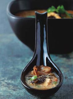 a black spoon with soup in it sitting on a table next to a bowl of food