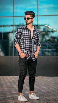 a man standing in front of a building wearing black pants and a plaid shirt with white sneakers