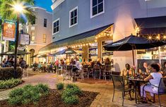 people sitting at tables in front of a restaurant with umbrellas and lights on the outside