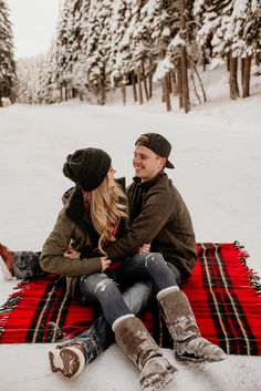 a man and woman sitting on top of a red blanket in the snow with their arms around each other