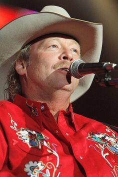 a man in a red shirt and cowboy hat singing into a microphone at a concert