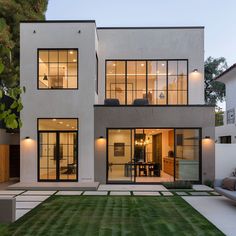 a modern house with large windows and grass in the front yard at dusk, as well as an outdoor seating area