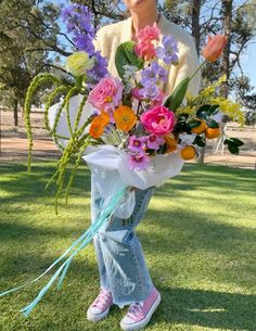 a woman holding a bouquet of flowers on top of a grass covered field with trees in the background