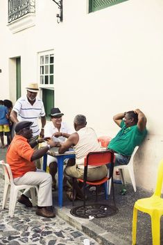 several men sitting at a table playing cards on the side of a building with yellow chairs