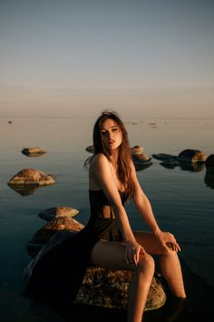 a woman sitting on rocks in the water with her legs crossed and looking off into the distance