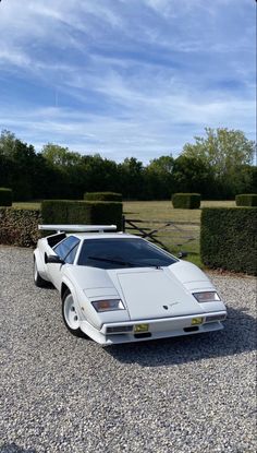 a white sports car parked in gravel next to hedges and bushes on a sunny day