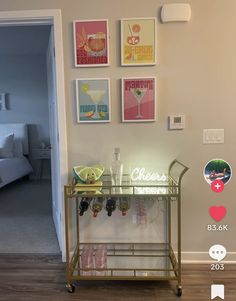 a bar cart in front of a wall with pictures on it and wine glasses sitting on the bottom shelf