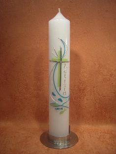 a white candle with a cross on the front and green leaves on the back, sitting against a tan wall