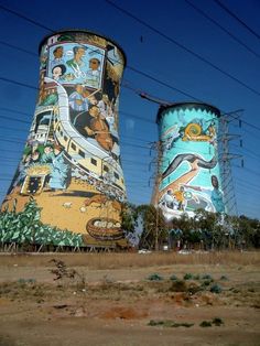 two large towers with paintings on them in the middle of a dirt field next to power lines