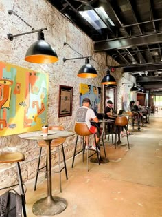 people sitting at tables in a restaurant with paintings on the wall and ceiling lights above them