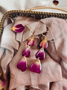 three purple flowers are hanging from gold earwires on a pink cloth in a basket