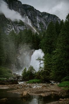 a waterfall in the middle of a forest filled with lots of trees and rocks under a cloudy sky