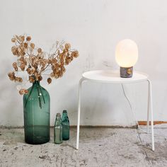 a green vase with dried flowers sitting next to two empty bottles on a white table