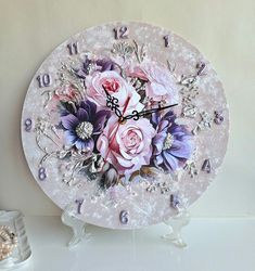 a white clock with pink roses and purple flowers on it's face sitting on a table